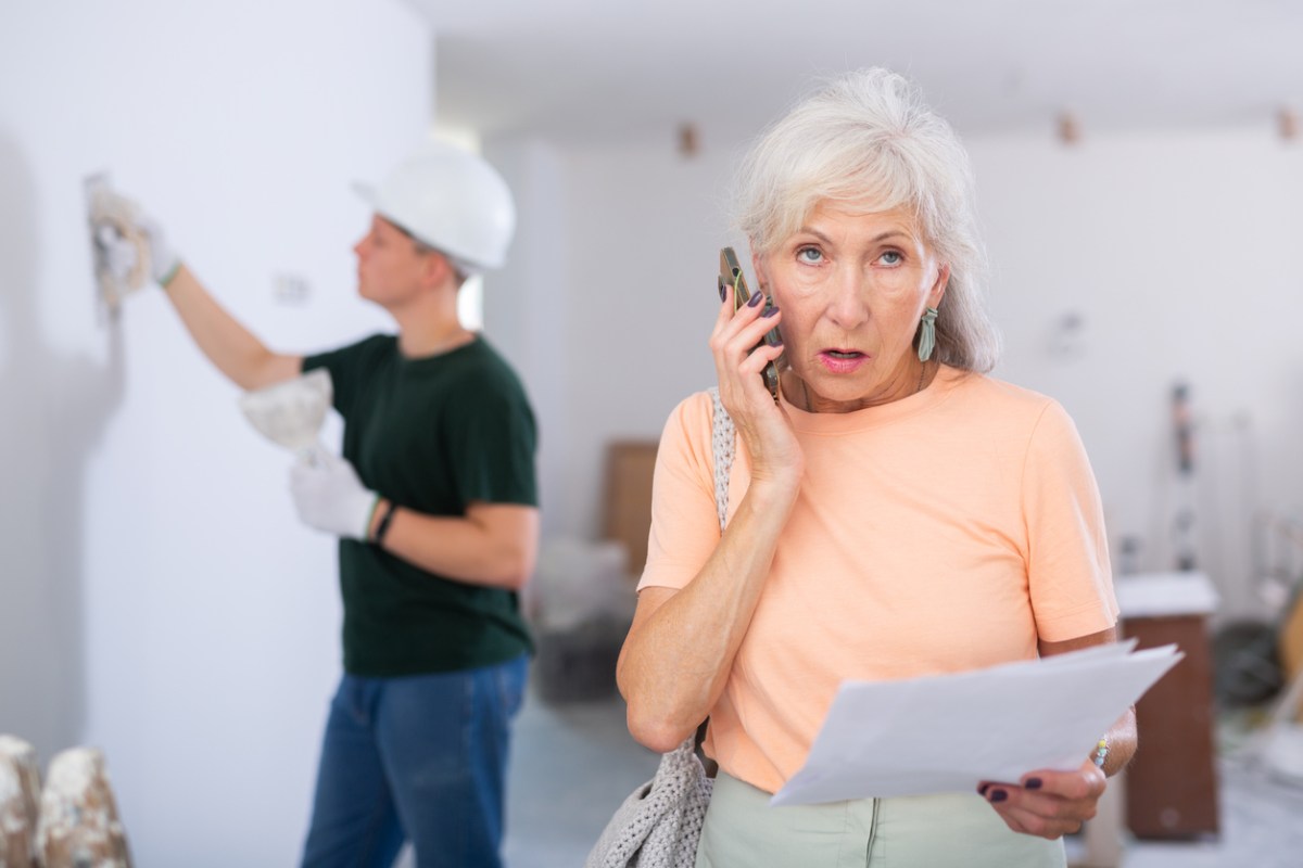 elderly woman in orange shirt talking on a cell phone while a contractor works on a white wall behind her