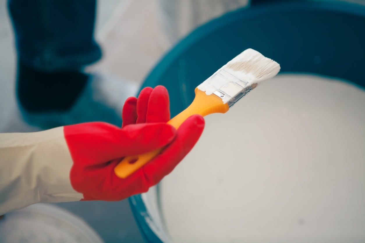 Close up of a person in a glove holding a paint brush over a bucket of paint