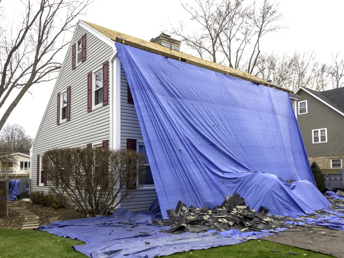 grey colonial home covered in large blue tarp with roof tiles on lawn while roof is being replaced