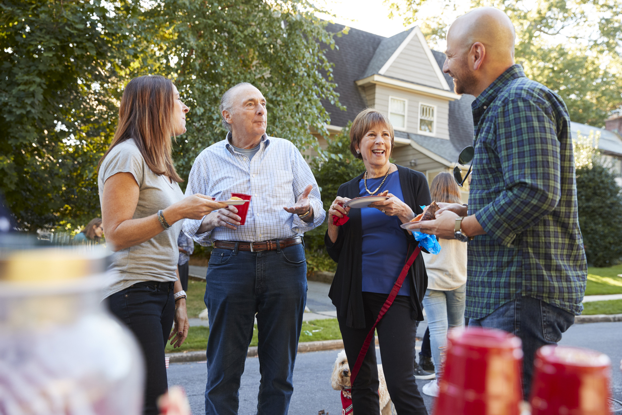 Middle aged and senior neighbours talking at a block party holding red plastic cups in front of large house