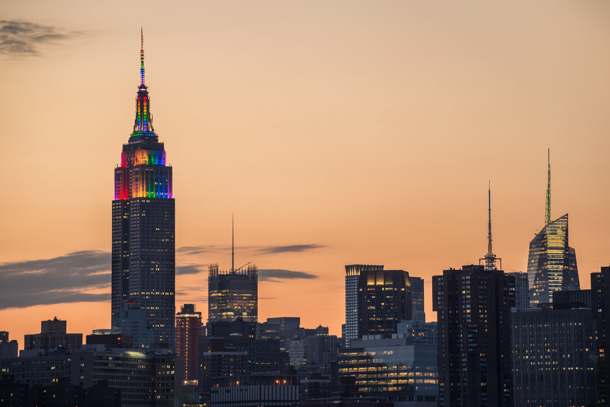 A partial skyline of Midtown Manhattan with the Empire State Building displaying Pride colors taken from Brooklyn just before at sunset.