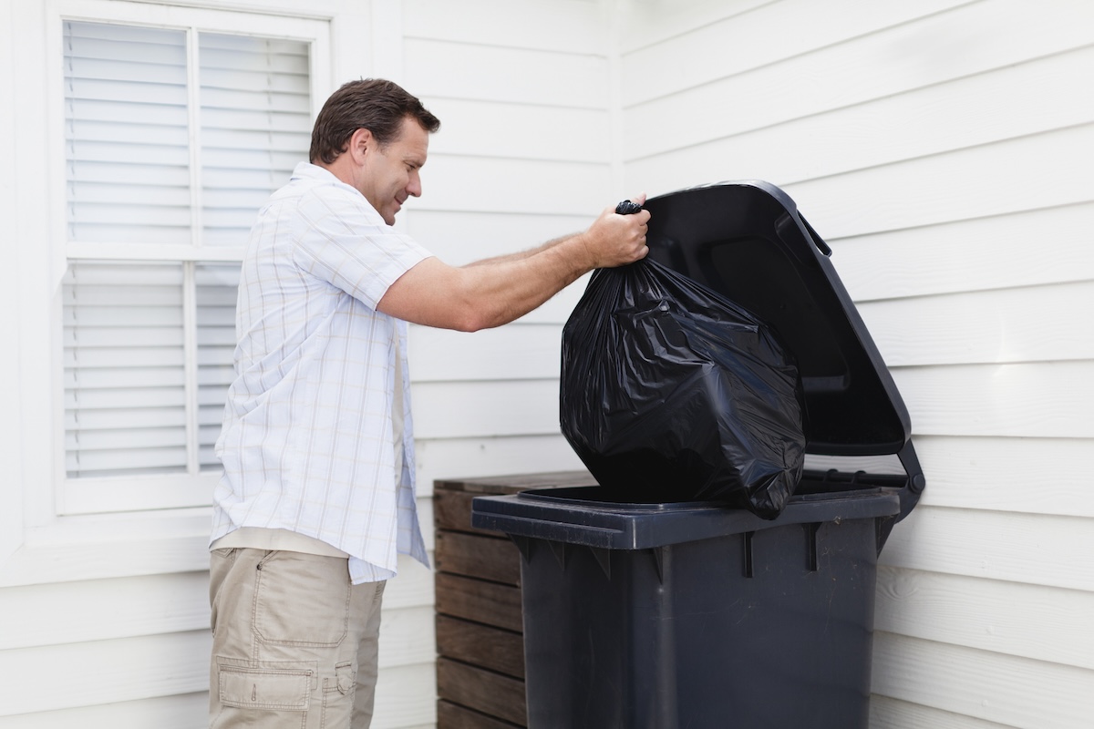 A man takes out a trash bag to an outdoor bin.