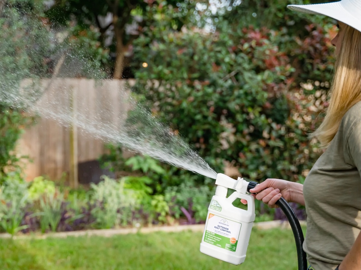 Woman wearing a wide-brimmed hat sprays her yard with Wondercide concentrate attached to garden hose.