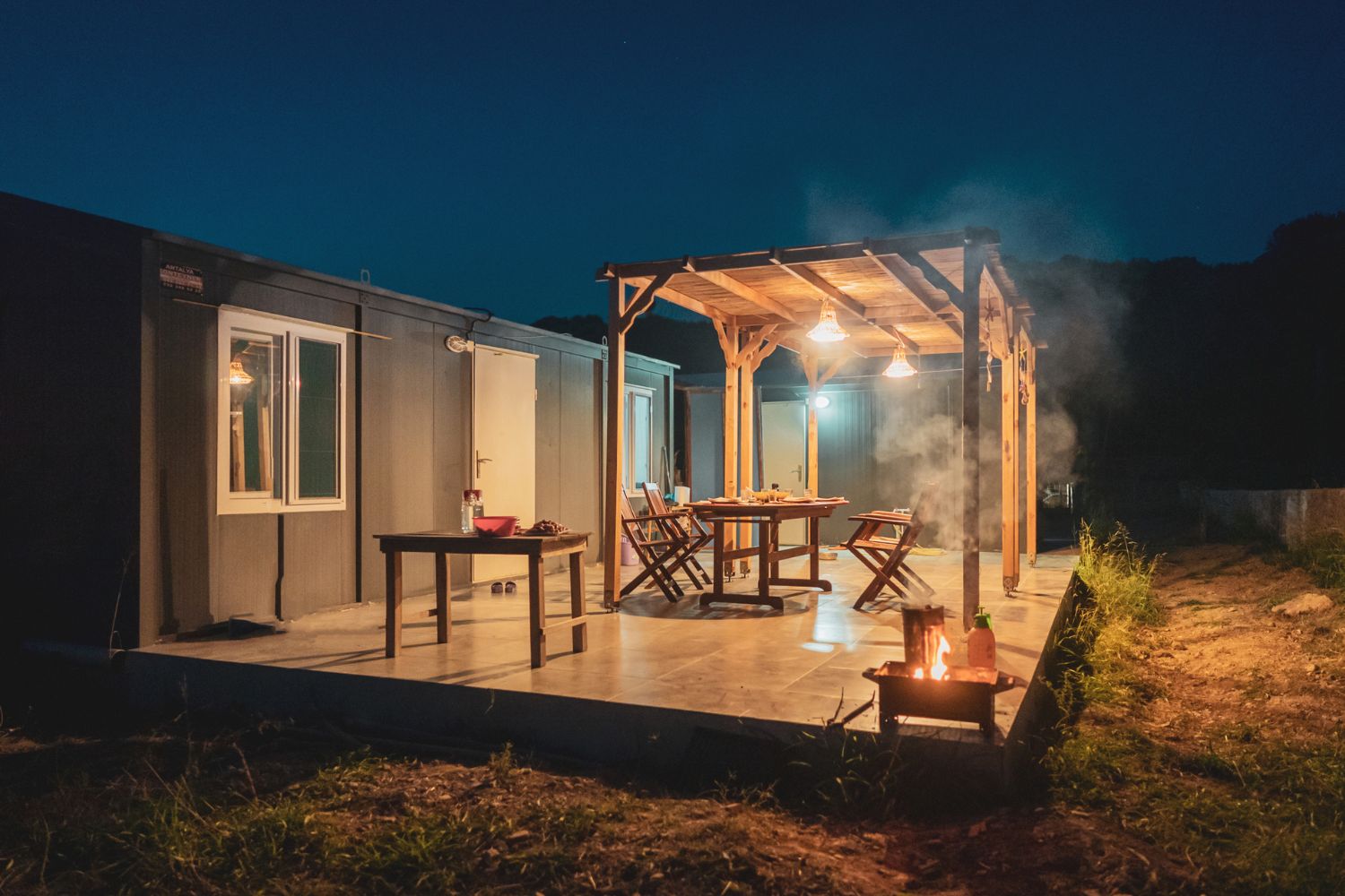 A nighttime view of the exterior of a container home. The back deck has a lit pergola, a table and chairs, and a fire pit.