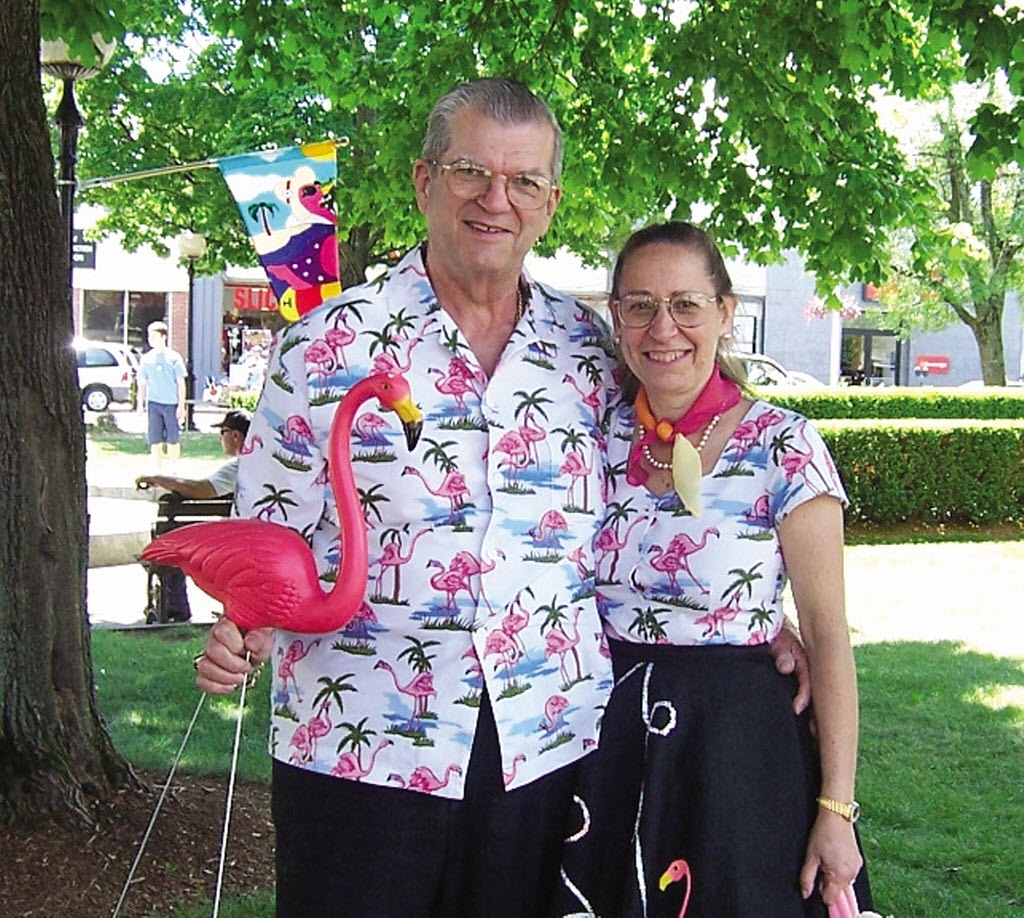 a senior man and woman in matching flamingo shirts stand outside under a tree holding a plastic flamingo