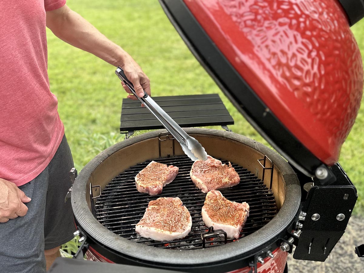 The Kamado Joe Classic Joe grill open while a person uses tongs to turn cooking meat