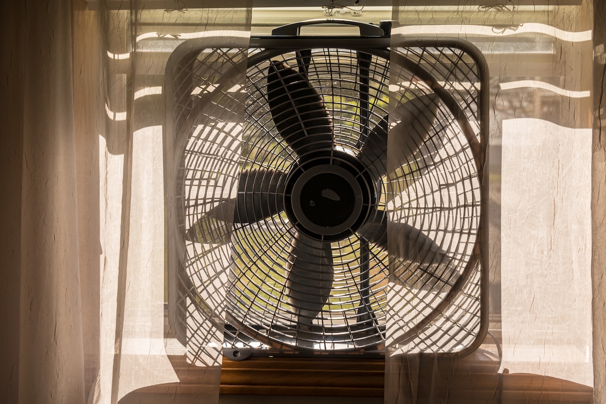 Electric box fan sitting on a window sill with sheer curtains in front of it.