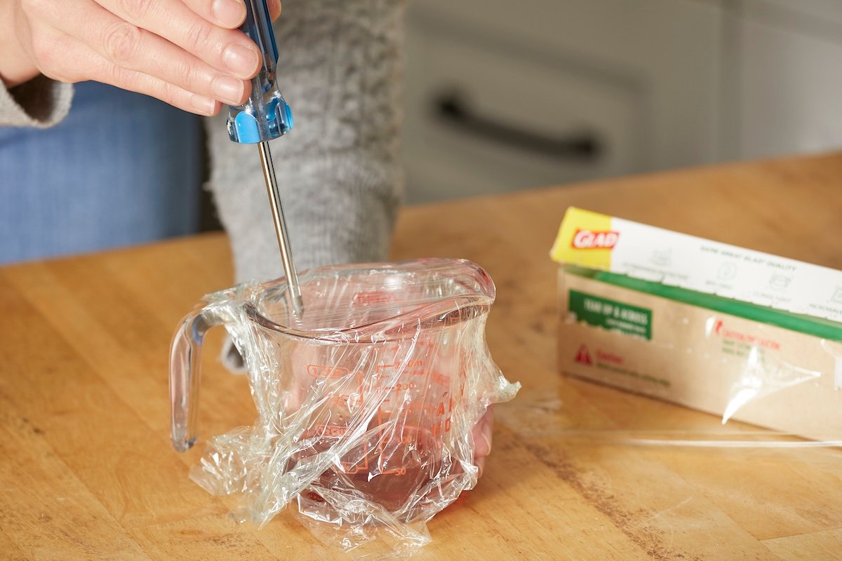 Woman uses a screwdriver to poke a hole through plastic wrap covering a measuring cup containing apple cider vinegar.