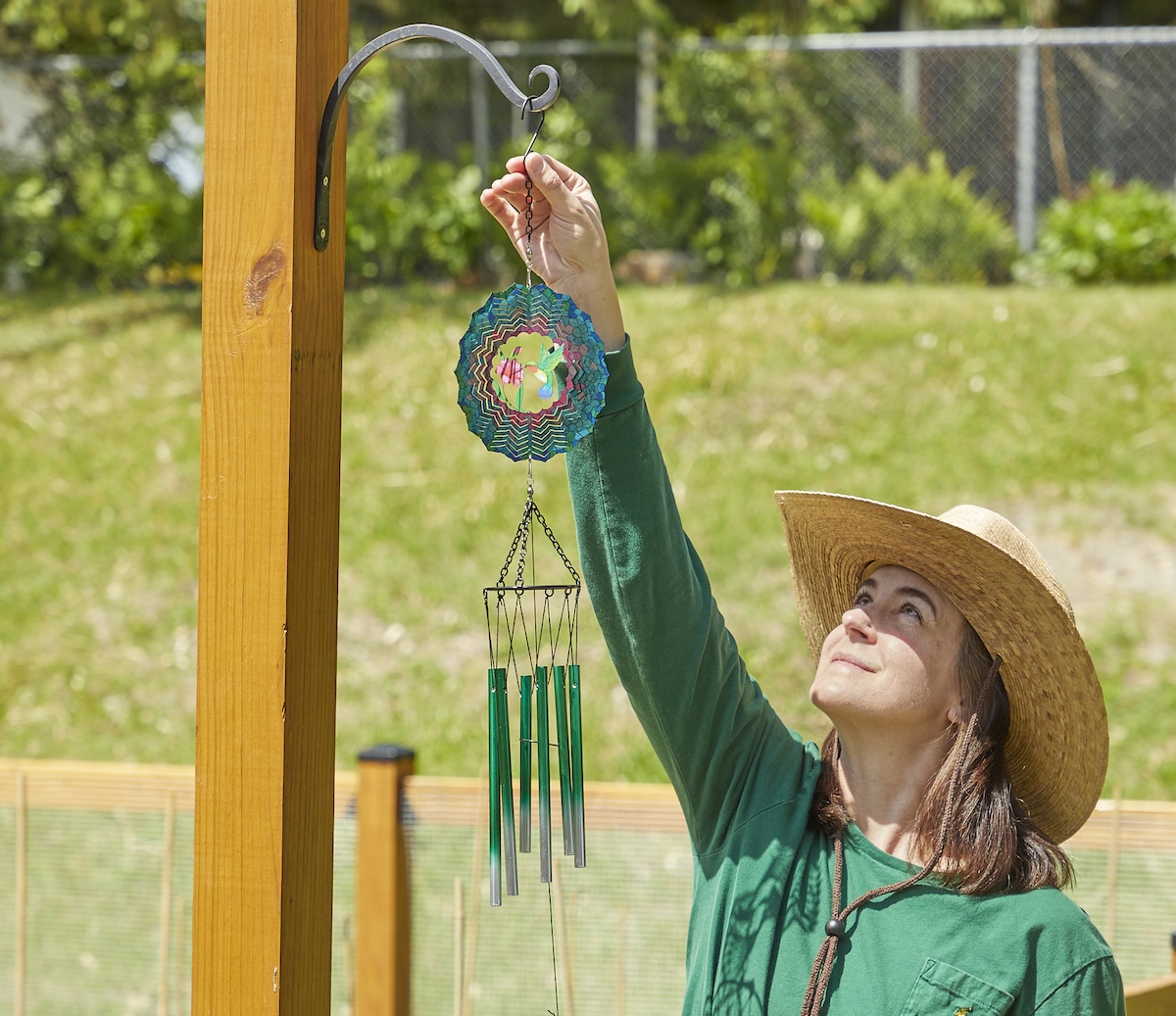 Woman wearing sun hat hangs wind chimes from a garden fence post.