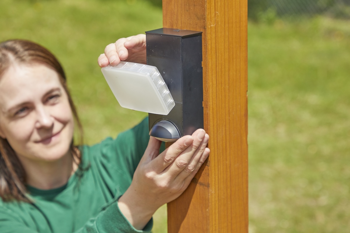 Woman hangs motion-sensing light on a wooden fencepost.
