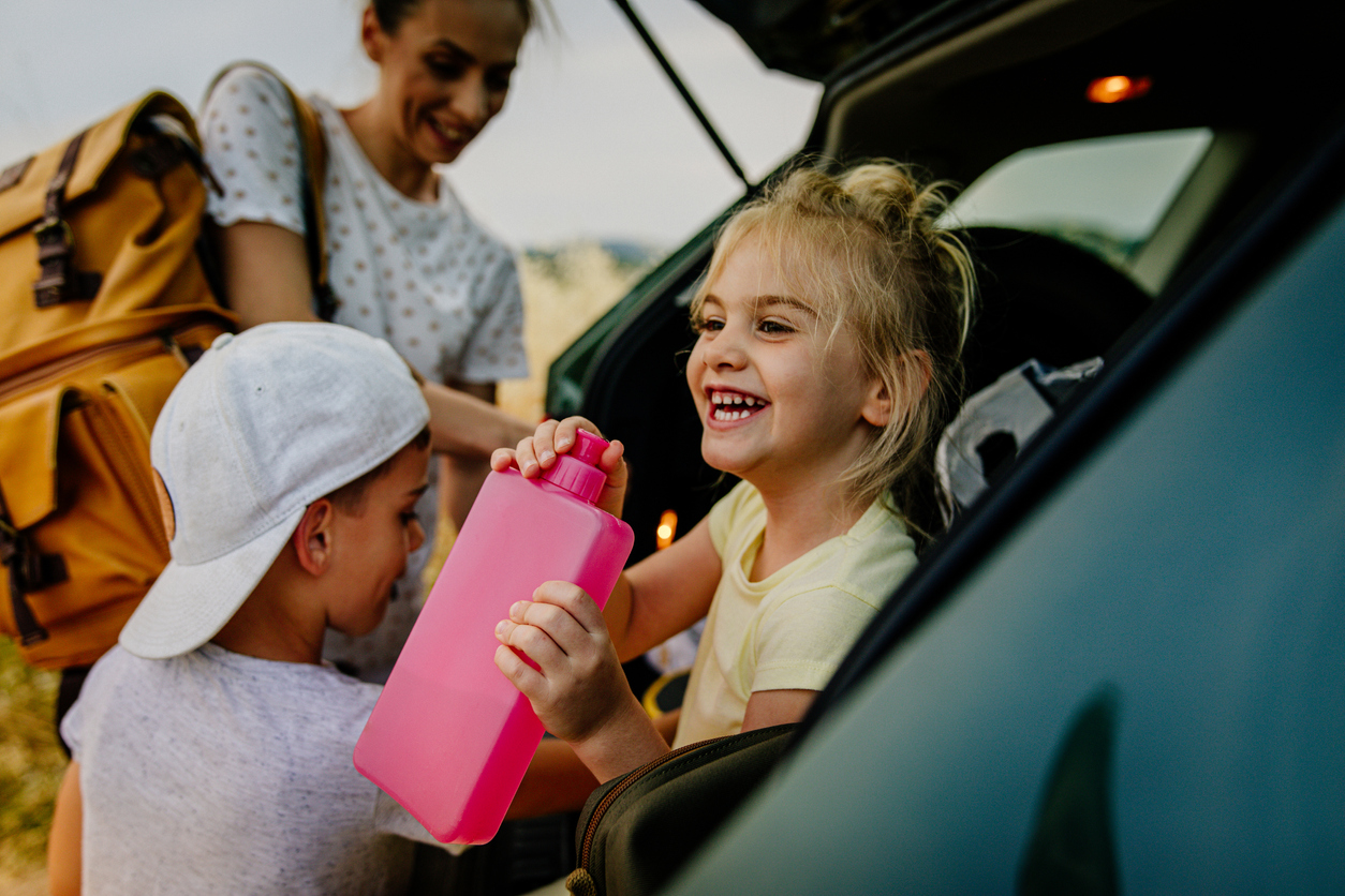 little girl sits in hatchback trunk drinking from pink water bottle as mother and brother load up the trunk for road trip