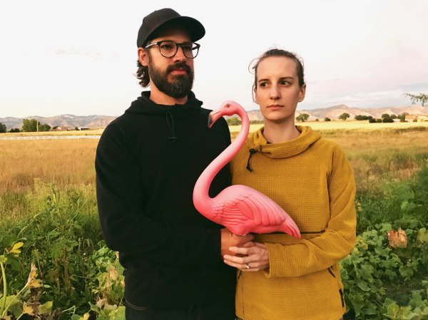 a-young-man-and-woman-in-a-field-hold-a-plastic-pink-flamingo-in-the-same-post-as-the-painting-American-Gothic