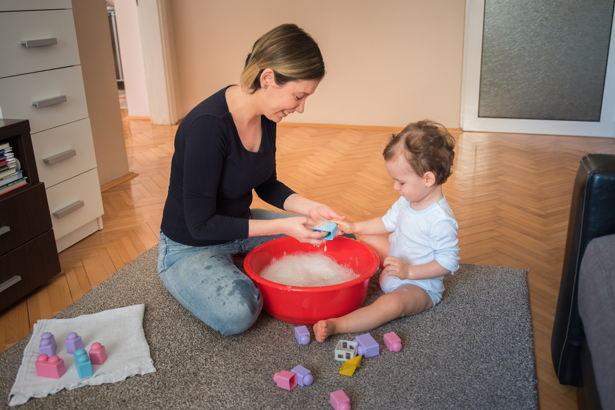 Young mother, woman, washing toys with her toddler son in a living room. Toddler playing with toys and water, making bubbles, enjoying his bonding time with mother. Coronavirus, covid 19 quarantine.