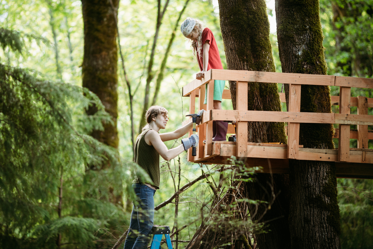 A woman and her child work together on the railings of a tree house in a large maple in their wooded back yard. A fun way to stay busy and follow social distancing.