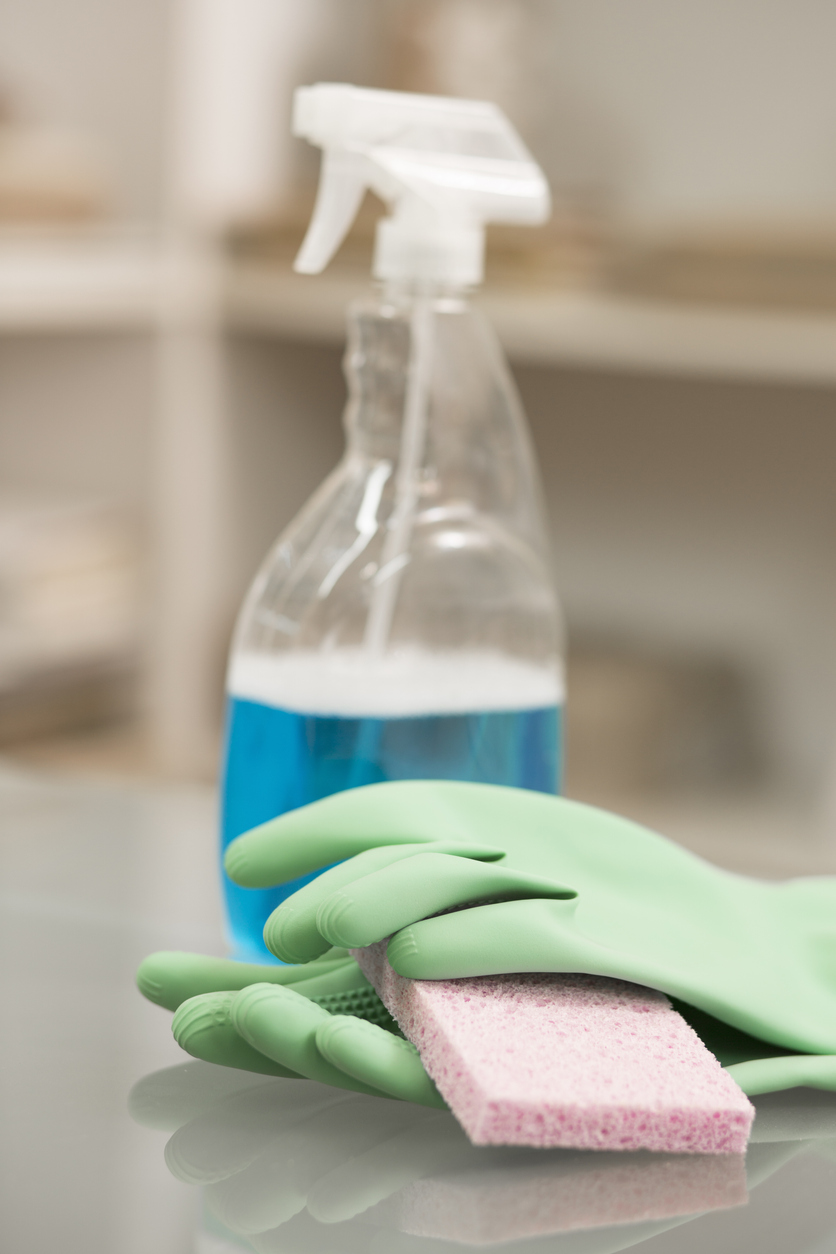 spray bottle with blue cleaning solution inside with gloves and sponge and blurry background on counter