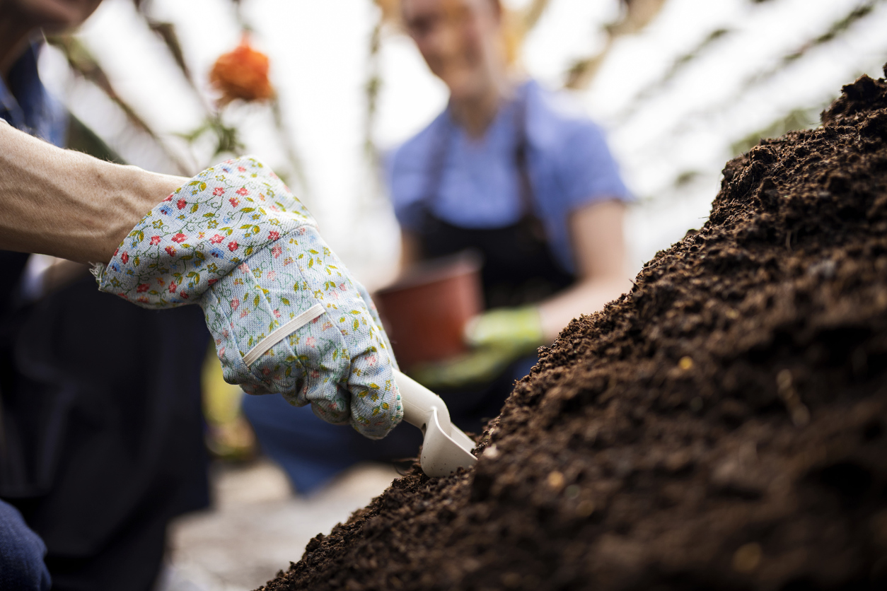 womans-hand-in-a-gardening-glove-scoops-soil-with-another-woman-in-the-background-holding-a-pot