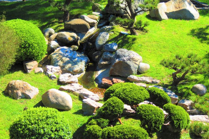 japanese garden with moss lawn and large rocks surrounding a small stream with bonsai trees near rocks
