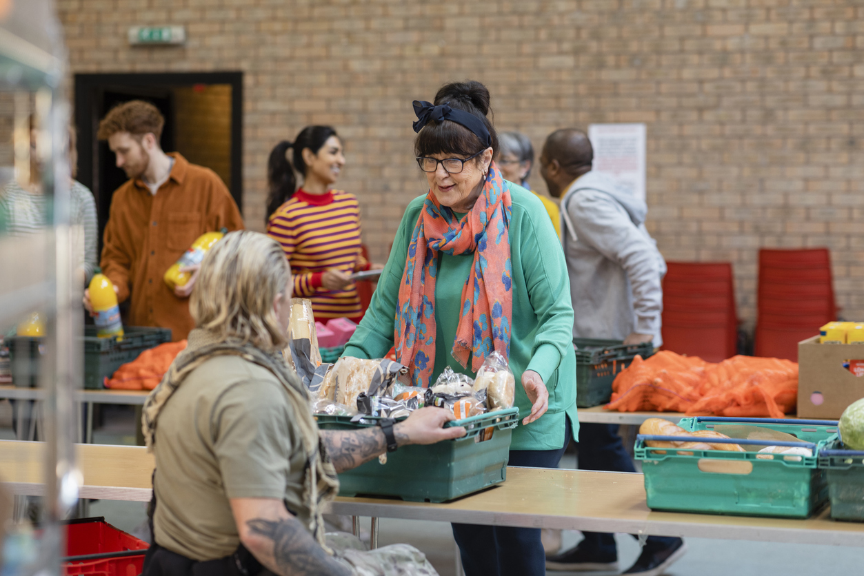 woman with glasses and bright orange scarf passes crate of food to another volunteer over a long table in a food bank with volunteers sorting food on tables