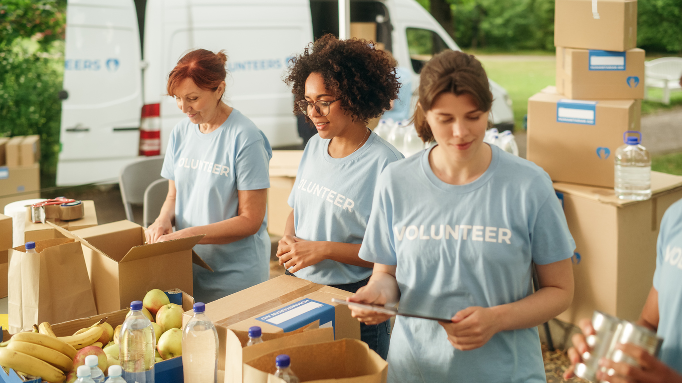 three-women-in-volunteer-tshirts-pack-food-into-cardboard-boxes-in-front-of-a-delivery-van