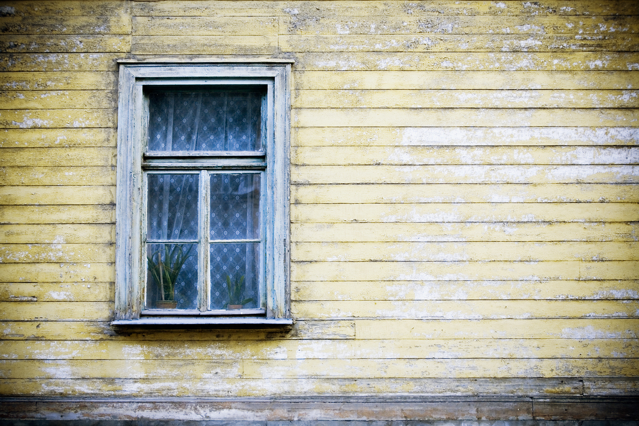 exterior wall of house with window with lace curtain and yellow siding that is faded and chipping