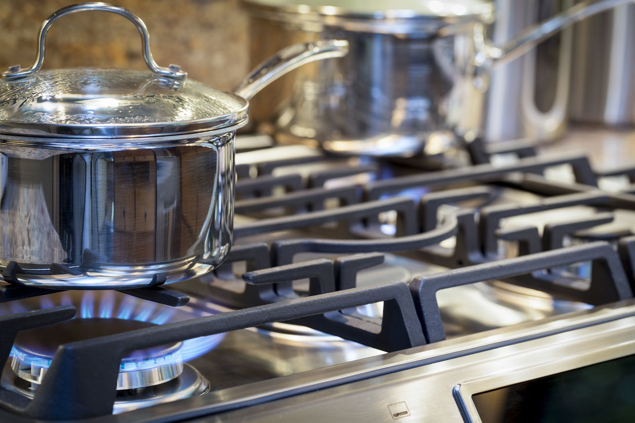 close shot of stainless steel pot with handle and glass lid on stove with gas on and another pot blurry in the background