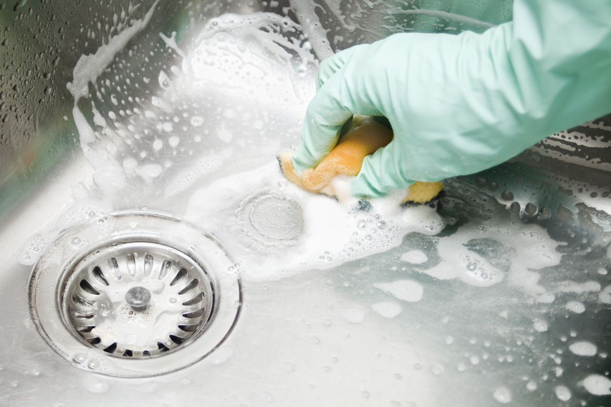 Human hands with protective gloves scrubbing up a square shaped large kitchen sink with a scouring pad, soap bubbles .