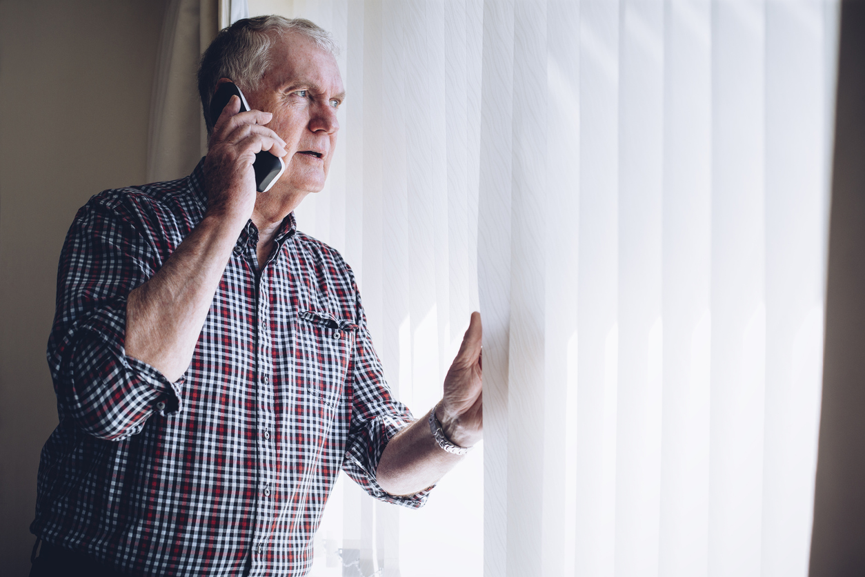 elderly man looking out window parting blinds and talking on cell phone