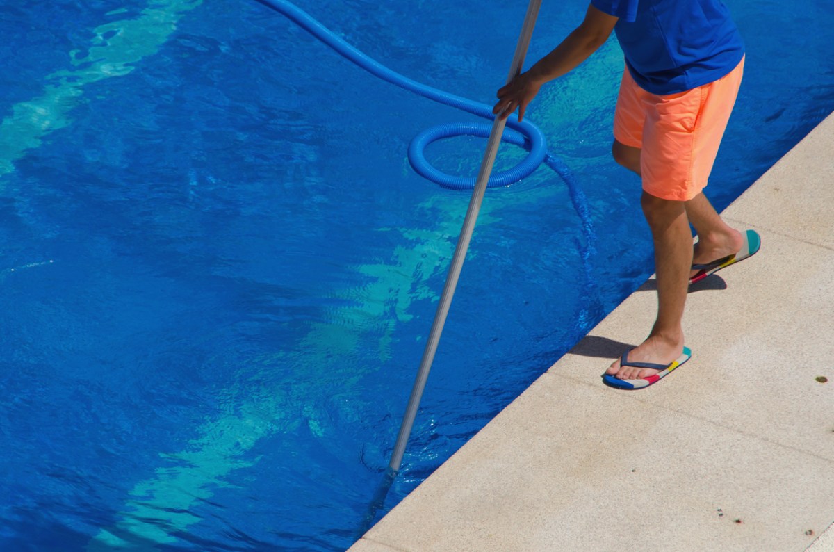 Man in orange shorts situates a pool vacuum in deep end of blue pool.