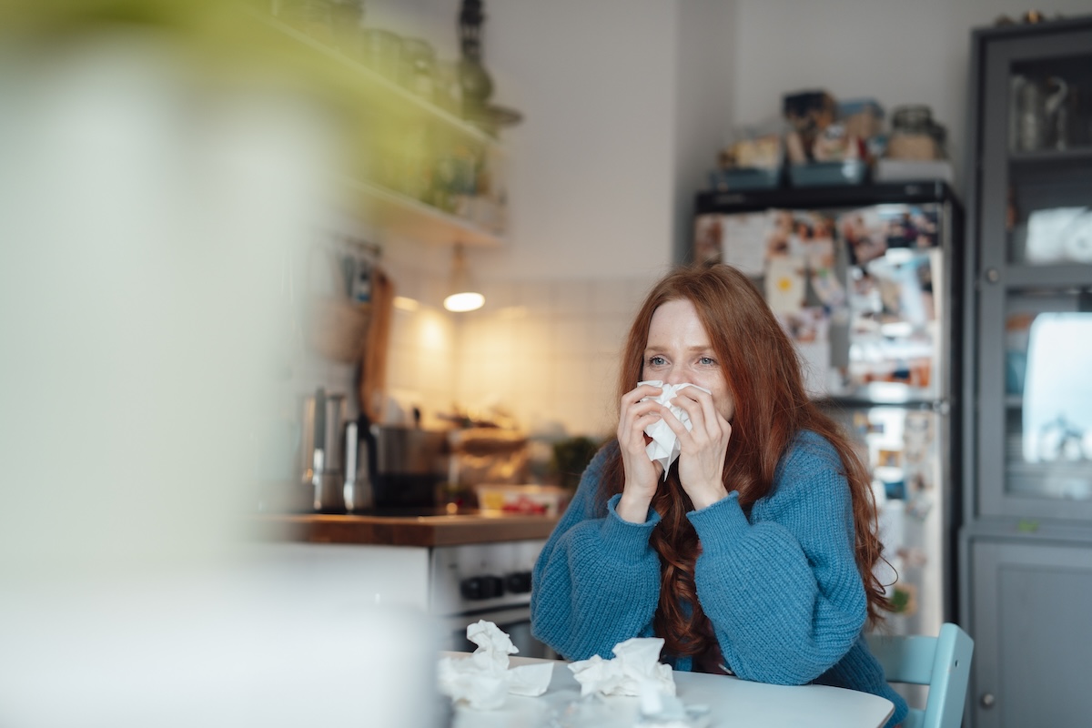 A woman with red hair sits at the kitchen table blowing her nose with tissues.