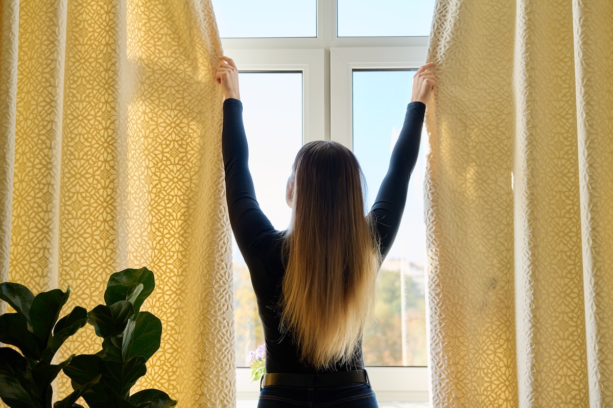 Young woman closing opening thick curtains on window, back view.
