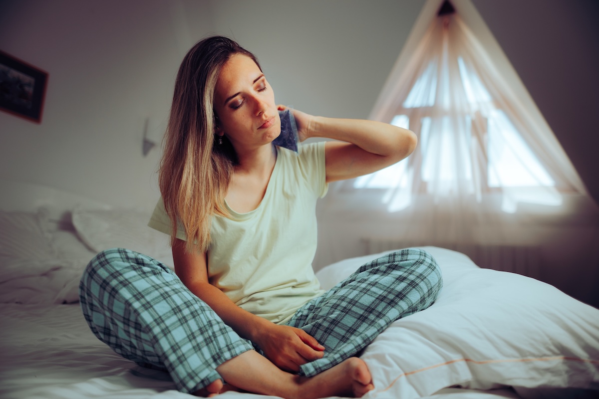 A young woman wearing flannel pajama bottoms and a T-shirt sits cross-legged on a bed, holding an ice pack to the back of her neck.