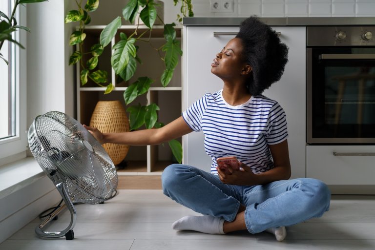 Young woman in striped T-shirt sits on floor with phone and tries to catch air currents emanating from fan.