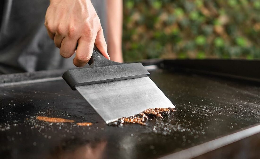 person cleaning a blackstone griddle with a metal scraper