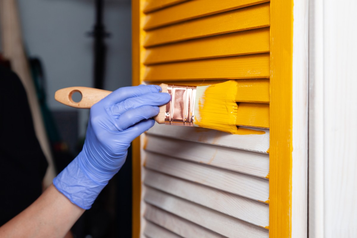 gloved hand using a paint brush with bright yellow paint on it and painting a white wooden window shudder yellow