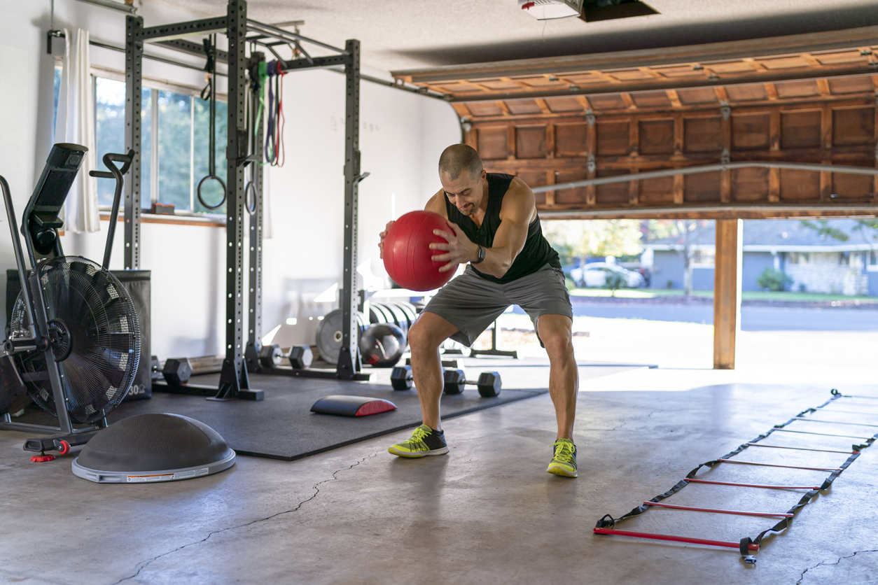A athletic man uses a medicine ball while doing a cross training workout in the gym he has set up in the garage of his home. He is staying healthy and active while isolating at home during the Covid-10 pandemic.