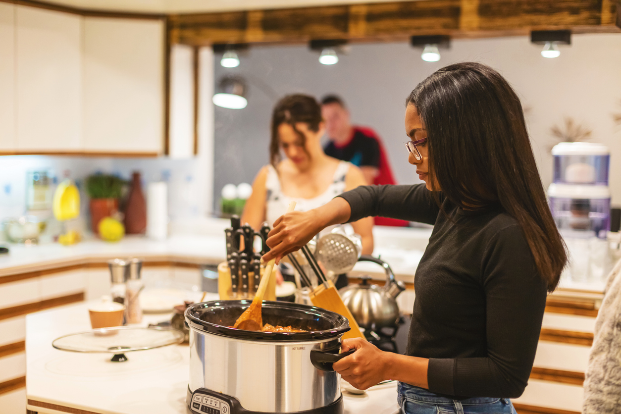 young woman stirring food in a slow cooker in a kitchen with other friends