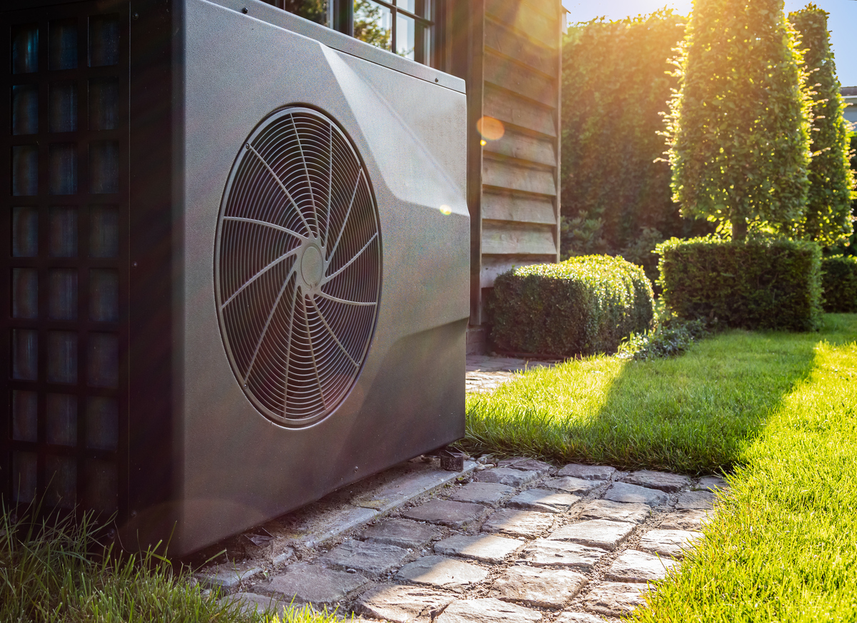 large heat pump with fan outside back of house with sunlit trees and shrubs