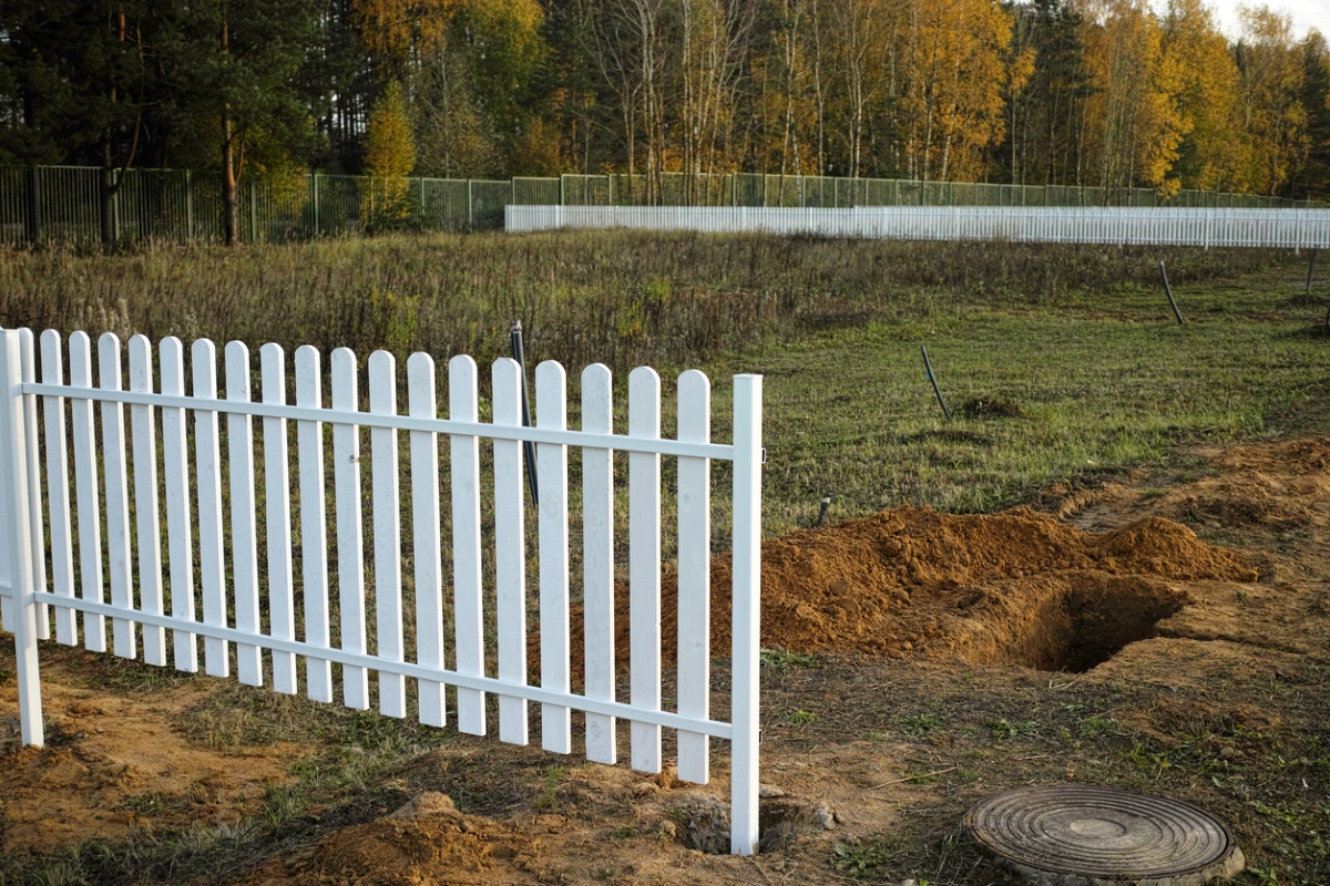 White picket fence near septic tank lid, with barren yard and dug holes in the background.