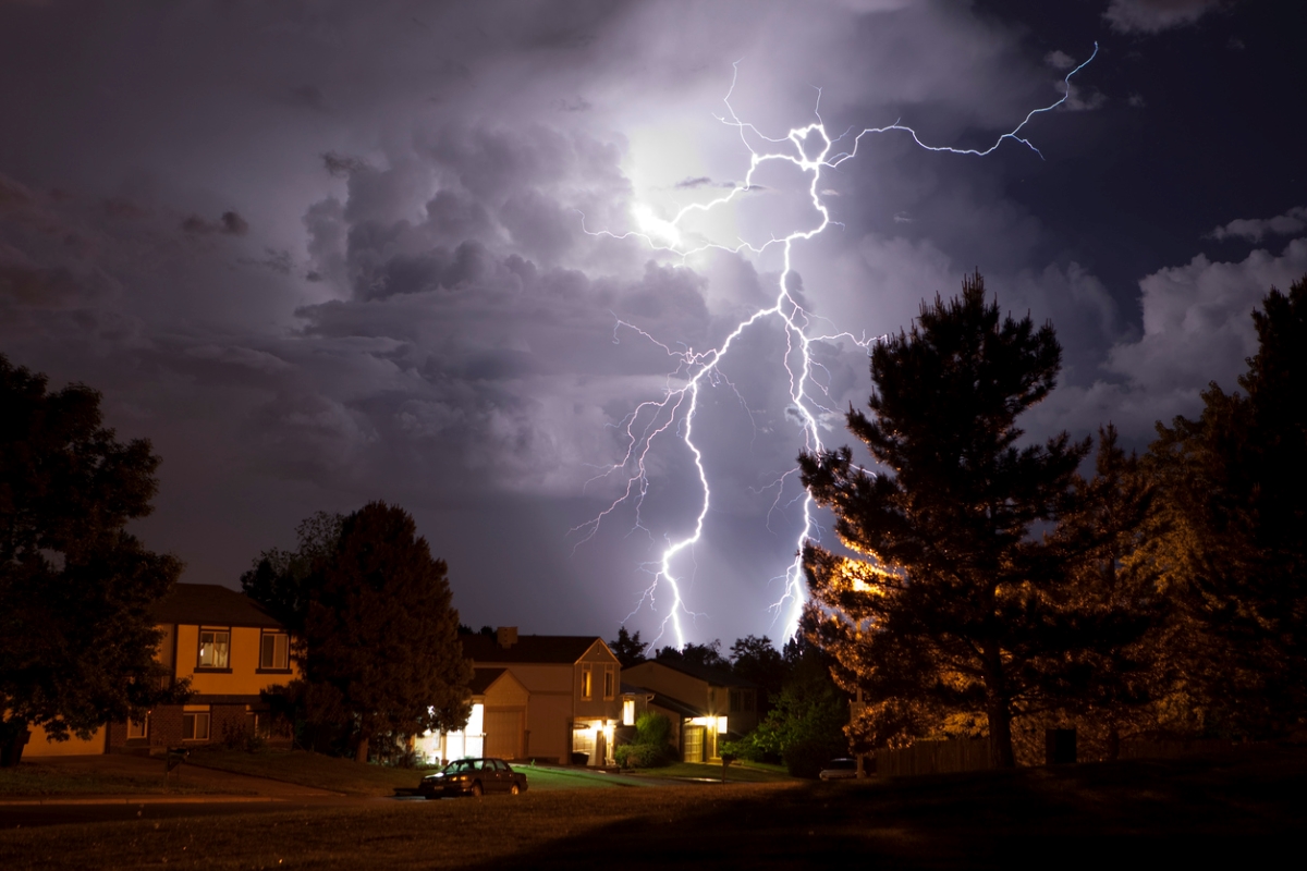 Lightening on dark night over an illuminated large tree and house.