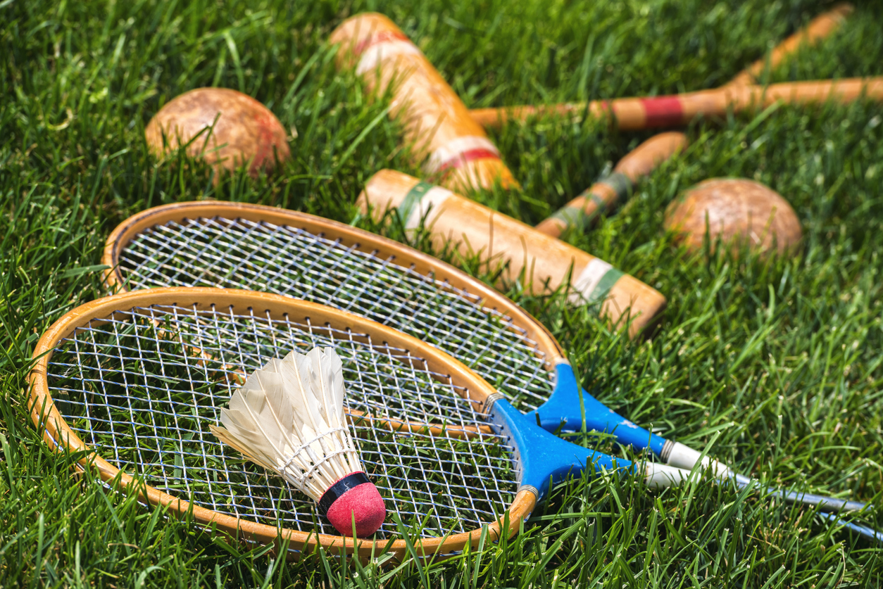 A pair of vintage wooden badminton rackets and a feathered shuttlecock lying in grass, along with a pair of croquet mallets and balls in the background. Great old fashion family fun games for all ages.