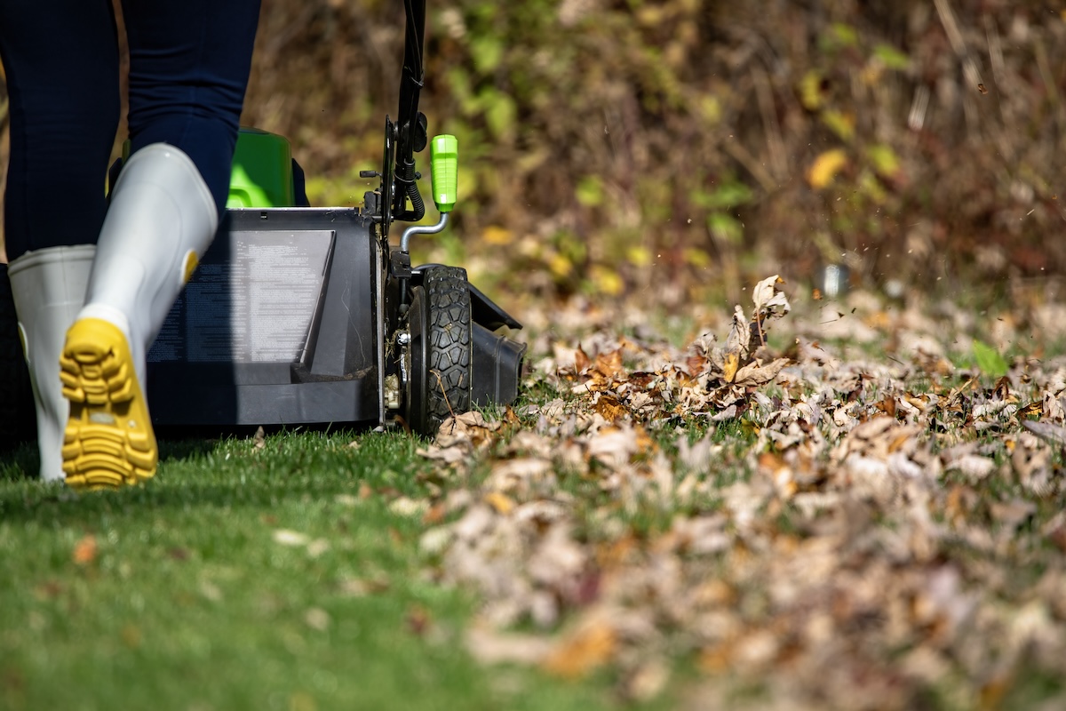A person wearing grey rain boots mows a lawn covered in a fall leaves.