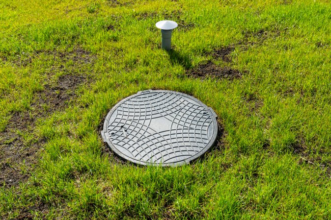 Septic tank cover and vent from a household septic tank, surrounded by patchy grass.