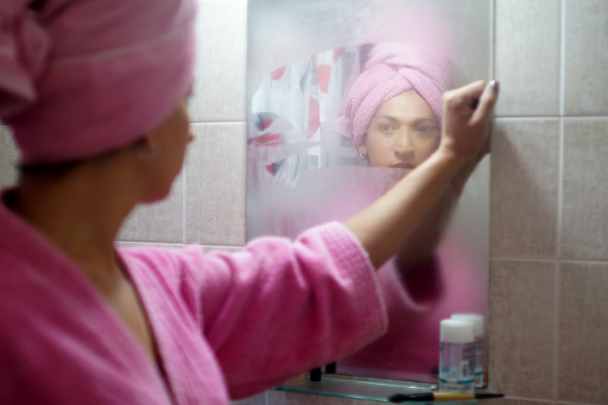 A women wearing a pink bathrobe and hair towel wipes steam from her bathroom mirror.