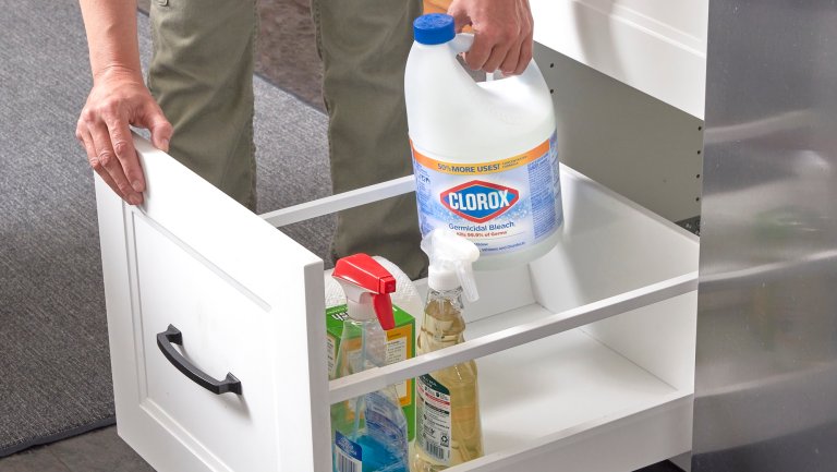 Woman puts gallon of bleach into a pull-out drawer with cleaning supplies.
