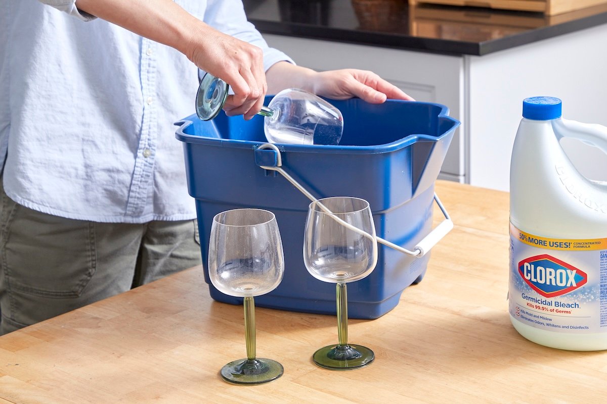 Woman dips wine glasses one by one into a blue bucket, a bottle of bleach nearby.