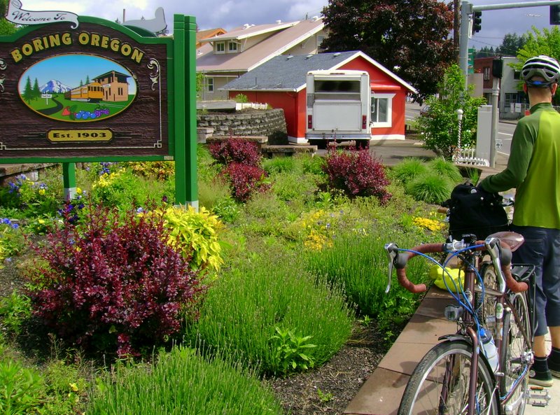 man with bike near sign for boring oregon with farmhouse in background