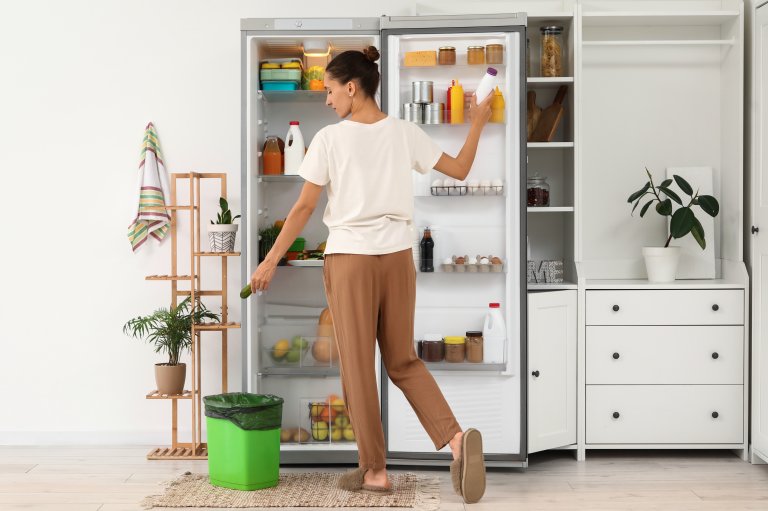 Young woman throwing spoiled food from open fridge in kitchen, back view