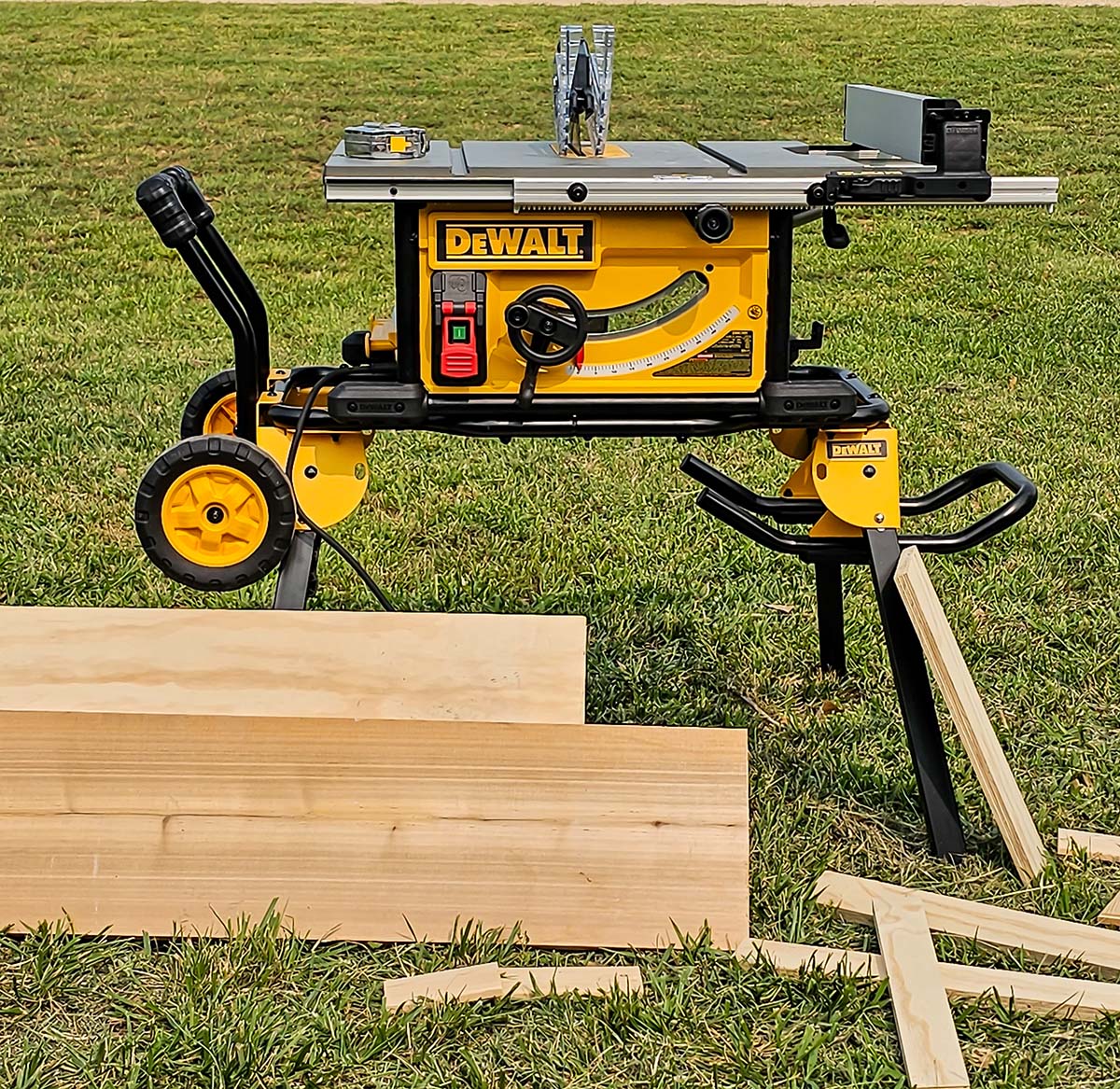 The DeWalt 10-inch table saw sitting on its stand in a yard while surrounded by several pieces of wood, some of which have been cut and some of which have not been cut