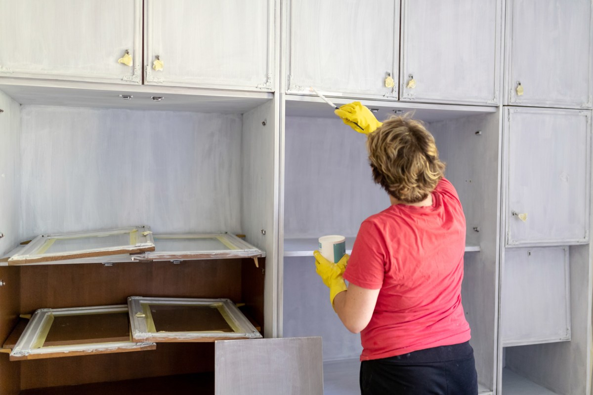 back of woman painting wall to wall cabinets white in diy project