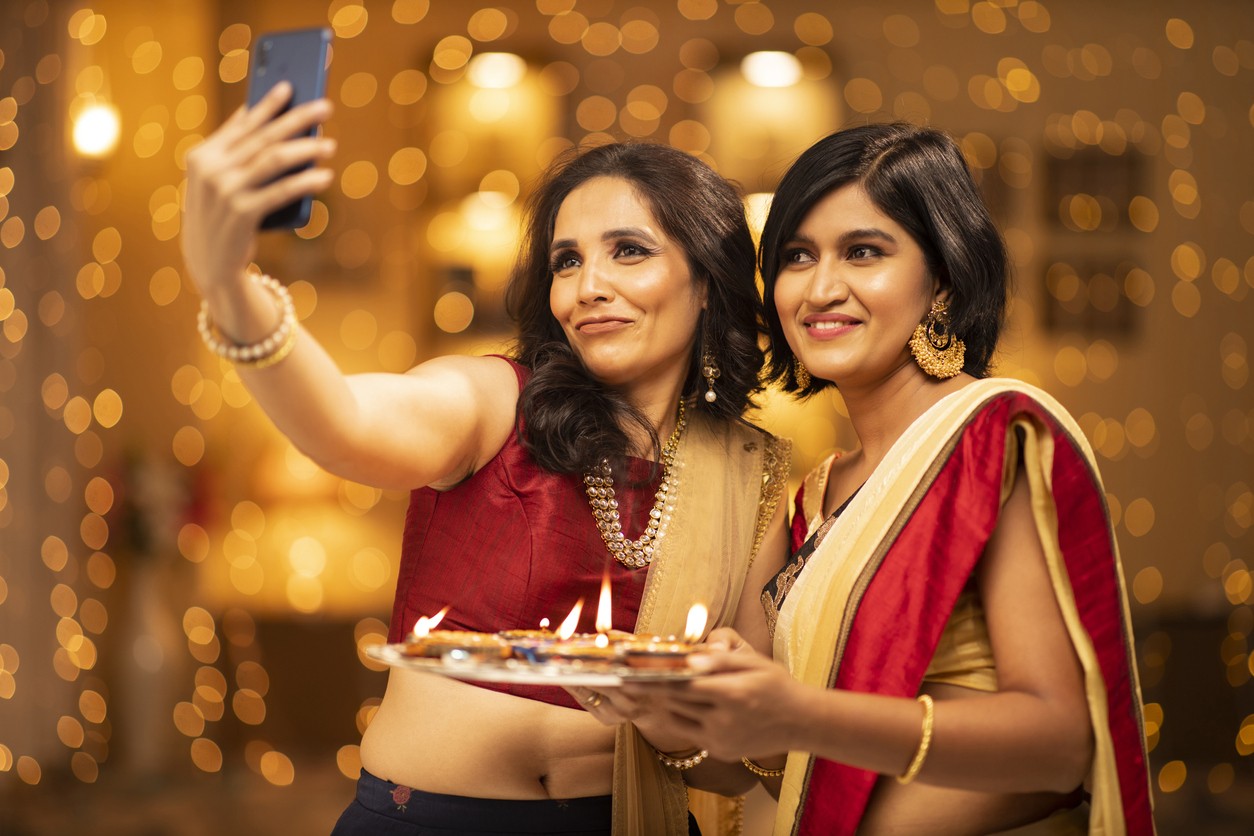 two-women-in-saris-take-a-selfie-in-front-of-string-curtain-lights-at-diwali-party